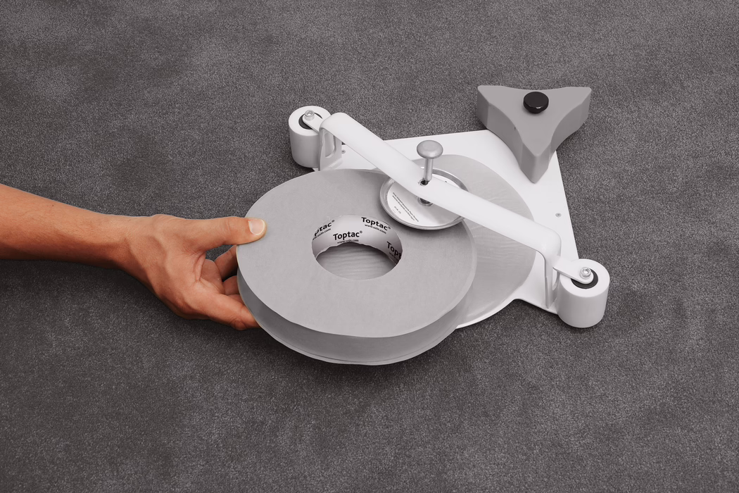 UZIN_Application_Image_Toptac 35_2_2018-03 Close-up of a left hand holding a large gray roll of adhesive tape positioned on a low-profile tape dispenser placed on a gray textured fabric surface. The dispenser features a white metal arm with a central spindle and a round cutting wheel, two small white rollers at the base and a triangular gray stabilizer with a black rubber foot; the cardboard core of the tape shows the printed brand 'Toptac'. Soft, even lighting emphasizes the neutral palette of grays and white. The composition centers on the tape roll and the hand in the lower left quadrant, showing skin tone and hand veins without any identifying features.