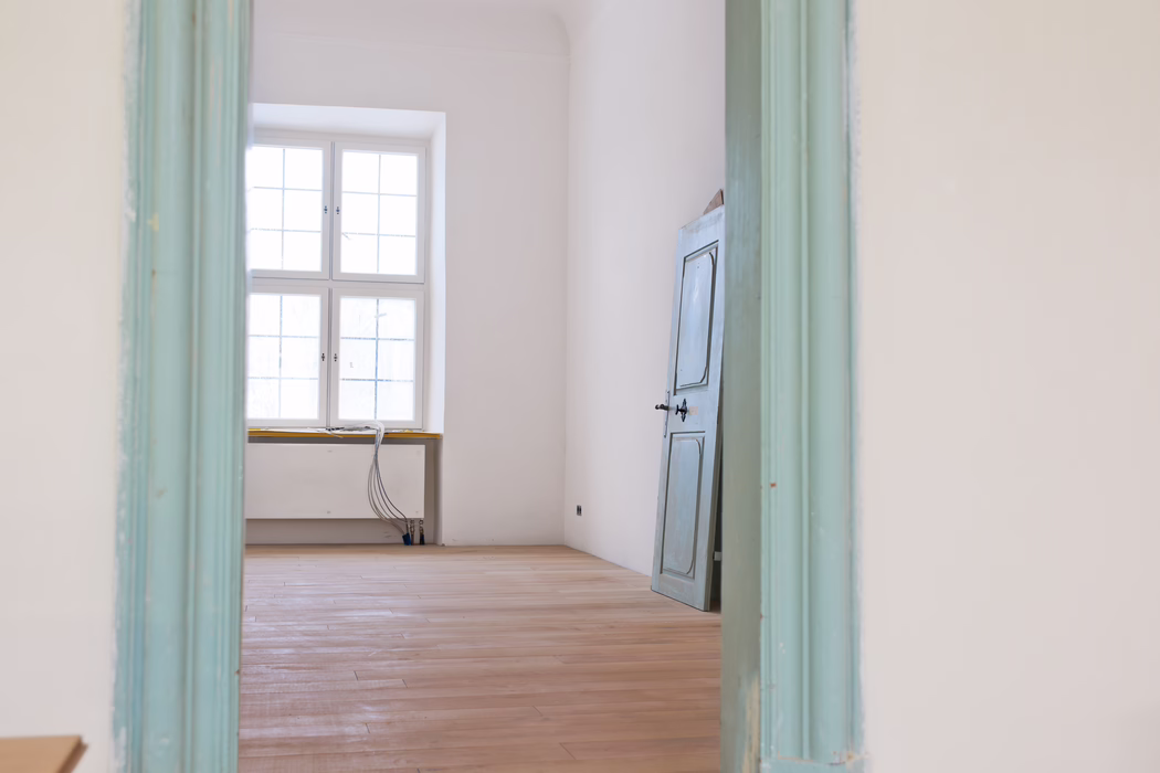 Interior view through a pale turquoise painted doorway into an empty, sunlit room. A large four-pane window centered on the far wall fills the space with soft natural light. The room has freshly sanded light wooden floorboards, white plaster walls and a detached, slightly weathered blue-gray wooden door leaning against the right wall. Near the window low heating pipes and a bundle of exposed electrical cables rest on the windowsill and descend to the floor, suggesting ongoing renovation work. The overall atmosphere is calm and minimal, with muted pastel tones and clean architectural lines.