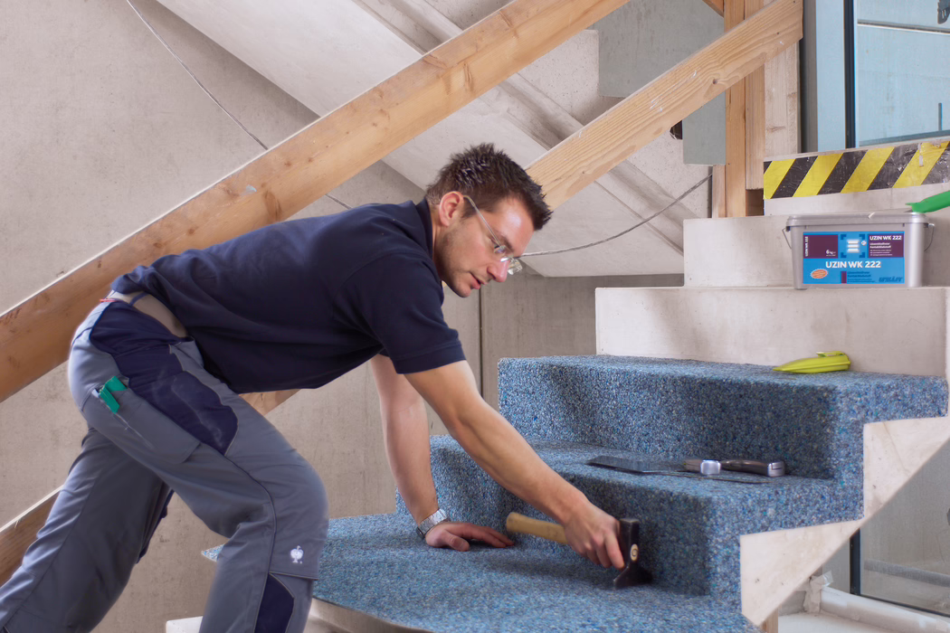 UZIN_Application_Image_WK 222_VA_4_ 2018-03 A construction worker is fitting a blue recycled carpet onto a set of unfinished concrete stairs inside a building under renovation. He is kneeling and leaning forward, wearing safety glasses, a navy polo shirt and grey work trousers, and using a rubber mallet to press the carpet firmly into place; nearby are a trowel, a tape measure and a plastic bucket labeled 'UZIN WK 222'. The scene is well lit with natural light, temporary wooden railing supports are visible, and the focus is on the careful, hands-on installation and the textured appearance of the carpet on the stair treads.