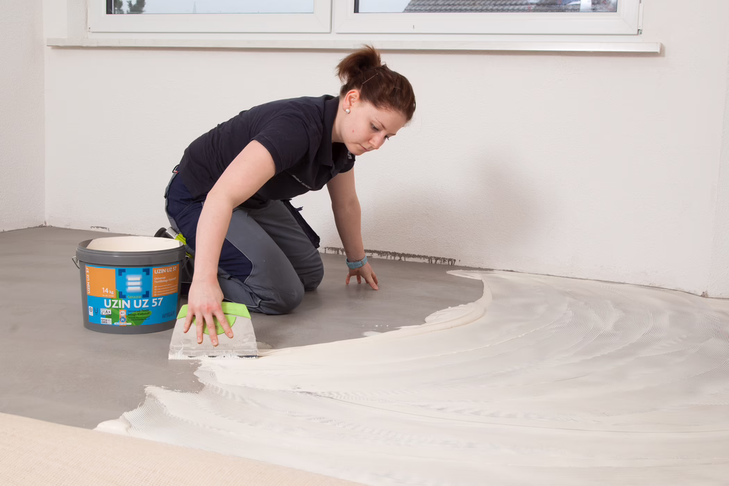 A person is kneeling on a bare concrete floor inside an unfurnished room, spreading a light-coloured adhesive or levelling compound with a wide notched trowel. A bucket labelled "UZIN UZ 57" stands nearby; the material is being applied in sweeping arcs toward the wall under a window. The worker wears dark work clothes, has their hair tied back, and focuses on creating an even layer for a subsequent floor covering.