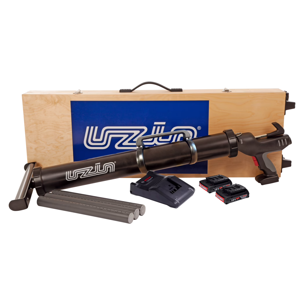 Wooden transport case with a black cordless caulking/adhesive gun and accessories: the top of a light plywood case with a black handle and a blue logo panel is visible behind a long black battery-powered caulking gun. In front of the case are three grey cartridge tubes, a battery charger base and three 18V lithium-ion battery packs. The gun features a pistol grip with grey and red accents, metal strap clamps around the cartridge barrel and a trigger assembly. Clean studio composition on a white background.
